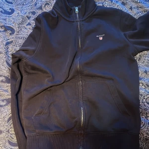 Svart ziptröja från GANT - Svart ziptröja från GANT med hög krage och dragkedja framtill. Tröjan har två stora fickor på magen och en liten GANT-logga broderad på bröstet. Ribbad mudd vid ärmslut och nederkant. Passar för dig som är 170cm