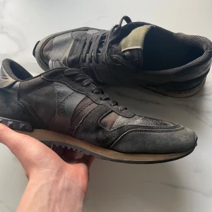 Valentino Rockrunner sneakers i svart - Säljer ett par svarta Valentino Rockrunner sneakers med kamouflagemönster och detaljer i skinn och mocka. Skorna har snörning, rund tå och en platt sula med nitar på hälen. Perfekta för dig som gillar exklusiva märken.