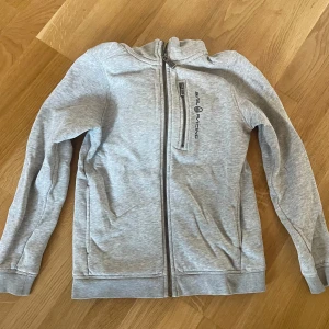Ljusgrå hoodie från Sail Racing i stl 140 - Säljer en ljusgrå hoodie från Sail Racing med dragkedja framtill och logga på bröstet. Tröjan har långa ärmar, huva och är i stl 140. Använt skick men har mer att ge, några mindre fläckar men som inte syns nämnvärt, se bilder. 