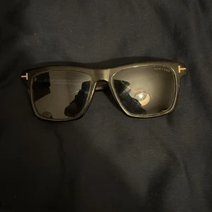 Svarta solglasögon från Tom Ford - Snygga svarta solglasögon från Tom Ford med klassisk rektangulär form och diskreta gulddetaljer vid bågarna. Glasögonen har mörka linser och en stilren design som passar till många tillfällen. Köpt för ca 3400 men använda lite, skriv privat för mer info/bilder.