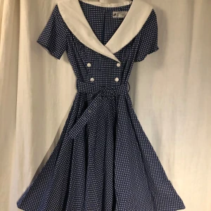 Blåvit prickig klänning från Hell Bunny Vixen - Retroinspirerad blå klänning med vita prickar från Hell Bunny Vixen. Klänningen har en stor vit krage, korta ärmar, knappar framtill och ett matchande midjeband. Perfekt för dig som gillar vintage-stil!