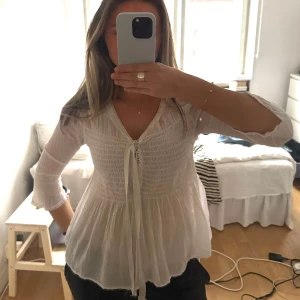 Vit blus från tessie - Supersöt och luftig blus i vitt från Tessie. Har smockad överdel som ger en fin passform och knytning framtill för en feminin touch. Vida ärmar med lätt volang i avslutet och ett fallande, vippigt tyg nedtill som gör den perfekt till både vardag och fest. Är v-ringad och har trekvartsärmar, i superbra skick🌟Står ingen storlek men uppskattas som en xs