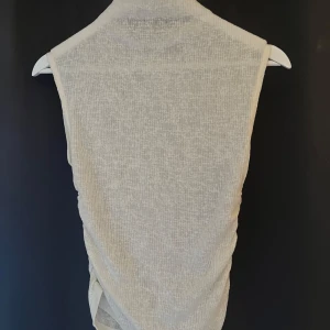 Vit ärmlös topp från Nelly - Säljer en vit ärmlös topp från Nelly i ett tunt, ribbat material. Toppen har en hög hals och lätt rynkade sidor för en snygg passform. Perfekt att styla med jeans eller kjol.