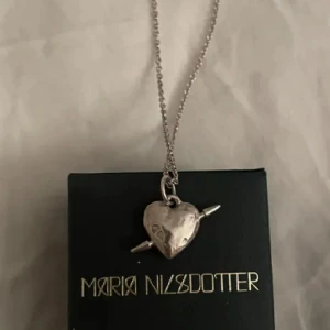 Silverfärgat halsband med hjärthänge från Maria Nilsdotter - Unikt halsband i silverfärg från Maria Nilsdotter med ett hjärthänge som har två spetsiga detaljer på sidorna. Kedjan är tunn och klassisk, perfekt för att ge en cool touch till din stil.