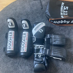Svarta boxningshandskar och benskydd från Booster - Säljer ett set med svarta boxningshandskar (12 oz) och benskydd från Booster Fight Gear. Handskarna har vita detaljer och kardborreknäppning. Perfekt för träning inom kampsport eller boxning. Knappt använt.