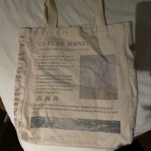 Beige tygväska med Claude Monet-tryck - Tygväska i beige med tryck inspirerat av Claude Monet. Väskan har två axelband och text samt bildmotiv på ena sidan. Perfekt för att bära böcker eller vardagliga saker. Materialet är kraftig bomullscanvas.