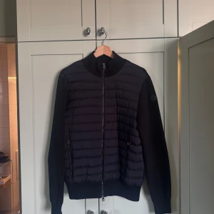 Svart pufferjacka från Moncler - Säljer en svart pufferjacka från Moncler med quiltade paneler framtill och släta ärmar. Jackan har dragkedja hela vägen, ribbade muddar och Moncler-logga på ärmen. Perfekt för dig som vill ha en stilren och sportig look.