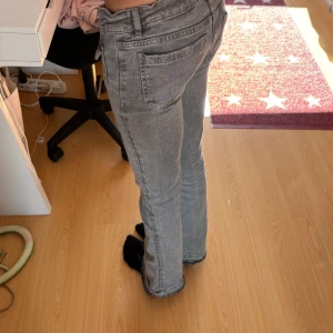 Grå bootcut jeans med fickdetaljer - Säljer ett par grå jeans med bootcut passform och snygga fickor framtill med knappar. Jeansen har låg midja och klassisk femficksdesign. Perfekta för dig som gillar en retro vibe och vill ha något som sticker ut lite extra.