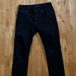 Svarta jeansbyxor från Nudie Jeans - Säljer ett par svarta slim jeans från Nudie Jeans med klassisk femficksdesign. Byxorna har midwaist och är tillverkade i ett slitstarkt jeanstyg. Storlek 30/32 modell ”Grim Tim” ny pris ca 1600. 