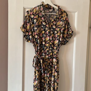 Blommig kortärmad klänning med knytband från Vero Moda - Säljer en blommig klänning från Vero Moda med korta ärmar och knytband i midjan. Klänningen har svart botten med gula, rosa och vita blommor. Perfekt för sommarens alla tillfällen!