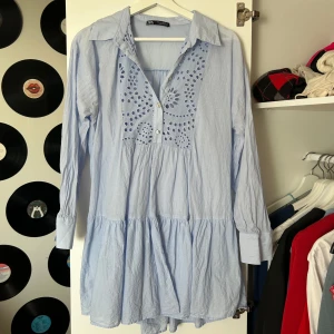 Ljusblå klänning med broderi - Ljusblå klänning från ZARA med lång ärm och v-ringning. Framsidan har fina broderade detaljer och knappar. Klänningen har en lös passform och volang nedtill, perfekt för en avslappnad och sommrig stil. 