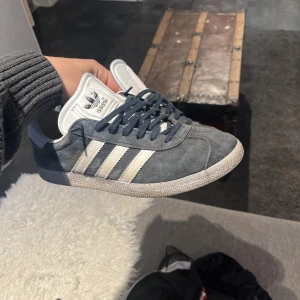 Adidas Gazelle grå/vita sneakers - Säljer ett par Adidas Gazelle sneakers i grå mocka med vita detaljer och klassiska tre ränder på sidan. Skorna har snörning, platt sula och rund tå. Perfekta för dig som gillar stilrena och tidlösa sneakers. Byten tas med.