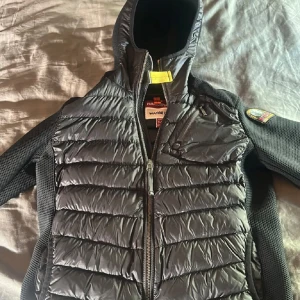 Svart dunväst med huva från Parajumpers - Säljer en svart dunväst från Parajumpers med huva och dragkedja framtill. Västen har quiltad framsida och rygg samt ett märke på ärmen. Snygg och sportig design med detaljer i mesh-tyg på sidorna.