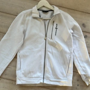 Vit jacka från Sail Racing med dragkedja - Säljer en vit jacka från Sail Racing med hel dragkedja framtill och ståkrage. Jackan har lång ärm, två sidofickor och Sail Racing-logga på bröstet. Perfekt för dig som gillar stilren och sportig design.