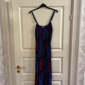 Sommarklänning - Säljer en långklänning med smala axelband och färgstarkt mönster i blått, lila och rosa. Klänningen har en lös passform och är perfekt för sommardagar. Materialet känns lätt och luftigt. Storlek är OneSize