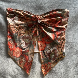 Blommig tubtopp med knyt - Säljer en snygg blommig tubtopp i beige, rött och grönt med knyt framtill. Toppen har ett unikt snitt med spetsig nederkant och är perfekt för sommaren. Mjukt och lätt material som känns skönt mot huden.