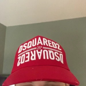 Röd keps från Dsquared2 - Snygg röd keps från Dsquared2 med vit broderad logga framtill och texten 'DEAN & DAN CATEN' baktill. Justerbart spänne i metall bak för perfekt passform. Perfekt accessoar för en sportig och trendig look.