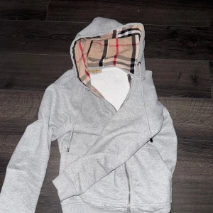 Grå hoodie med dragkedja från Burberry - Säljer en grå hoodie från Burberry med klassiskt rutigt foder i huvan. Tröjan har dragkedja framtill, fickor på sidorna och justerbar huva med snören. Perfekt för en avslappnad och stilren look.