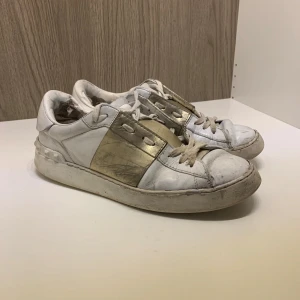 Vita sneakers med gulddetalj från Valentino - Säljer ett par vita sneakers från Valentino med en bred guldfärgad rem över sidan. Skorna har rund tå och platt sula, samt klassisk snörning framtill. Perfekta för dig som gillar stilrena skor med en touch av lyx.