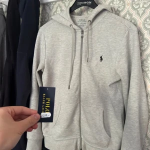 Ljusgrå hoodie från Polo Ralph Lauren - Säljer en stilren ljusgrå hoodie från Polo Ralph Lauren med dragkedja och huva. Tröjan har två fickor framtill och den klassiska broderade loggan på bröstet. Perfekt för en avslappnad look.