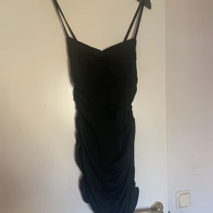 Svart tight klänning från Offordram - Säljer en svart tight klänning från Offordram med smala axelband och rynkade detaljer framtill. Klänningen är tillverkad i stretchig polyesterblandning och har en figurnära passform som framhäver siluetten. Perfekt för dig som vill ha en stilren look.