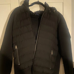 Svart Cardigan Från Moncler - Säljer en svart Cardigan från Moncler med stickade ärmar och huva. Jackan har två snedställda fickor med dragkedja framtill och en Moncler-logga på ärmen. Perfekt för kyliga dagar och riktigt stilrent. Pris 850 kr eller billigare vid snabb förhandling