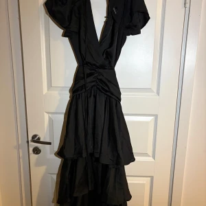 Balklänning - Säljer en svart långklänning från Boohoo med v-ringning och volangdetaljer. Klänningen har korta ärmar och omlottmodell i midjan som ger en snygg siluett. Perfekt för dig som vill ha en elegant klänning till balen. Nyskick med prislsppen kvar