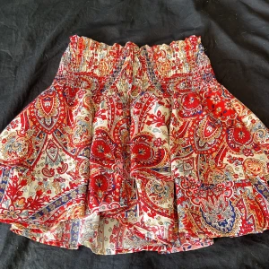 Röd- och vitmönstrad kortkjol med smock - Supersöt kortkjol med färgstarkt paisleymönster i rött, blått och beige. Kjolen har bred smock i midjan med knytband framtill och inbyggda shorts. Perfekt för dig som gillar boho-stil och vill ha något luftigt och bekvämt.