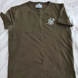 Olivgrön t-shirt från SikSilk stl S - Olivgrön t-shirt från SikSilk med vit broderad logga på bröstet. Klassisk rund halsringning och kort ärm. Tillverkad i mjuk bomull för en skön känsla.