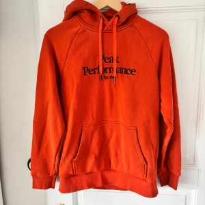 Orange hoodie från Peak Performance - Säljer en orange hoodie från Peak Performance med broderad logga på bröstet. Tröjan har huva med dragsko och en stor magficka. Perfekt för dig som gillar en sportig och avslappnad stil.