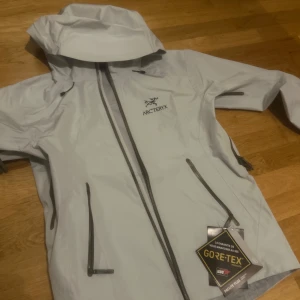 Vit skaljacka från Arc'teryx beta lt - Säljer en Vit Arc'teryx jacks med huva och vattentätt GORE-TEX-material. Jackan har dragkedjor framtill och på fickorna, samt diskret logga på bröstet. Perfekt för utomhusaktiviteter när vädret är ostadigt.