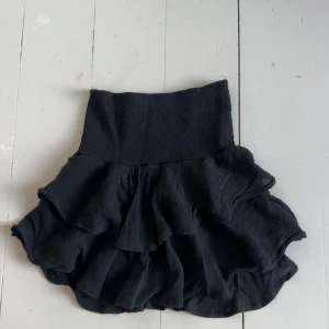 Svart volangkjol - Supersnygg svart kjol med flera volanger. Kjolen har en kort längd och är perfekt för dig som vill ha en trendig och feminin look. Materialet är lätt och ger ett fint fall. Använt fåtal gånger.