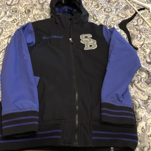 Svart och blå jacka från Special Blend - Cool svart jacka med blå ärmar från Special Blend. Jackan har huva, dragkedja framtill och broderad SB-logga på bröstet. Insidan har ett unikt mönster med text och bilder. Perfekt för dig som vill sticka ut. Unik jacka har använt för skidåkning men är alldeles för liten nu 