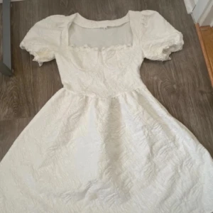Vit klänning med puffärmar - Superfin vit klänning med puffiga kortärmar och fyrkantig urringning. Klänningen har en lätt struktur i tyget och en utställd kjol. Perfekt till studenten, skolavslutning eller en fin sommardag!