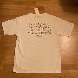 Vit t-shirt från Maison Margiela - Vit t-shirt från Maison Margiela med tryck av siffror och logga på ryggen. Klassisk passform med korta ärmar och rund hals. Tillverkad i mjuk bomull, perfekt för en clean och stilren look. Renade i garderob och hitta denna, försöker bli av med den så jag kan känna ihop en slant till sommaren