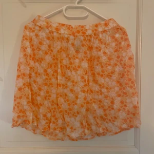 Blommig kjol från Pieces - Säljer en söt kortkjol från Pieces med blommigt mönster i orange och vitt. Kjolensbaksida har resår i midjan och är lätt och luftig, perfekt för varma dagar. Passar dig som gillar färg och mönster! Storlek M/38💖använt 1-3 gånger och är bara lite skrynklig 💖