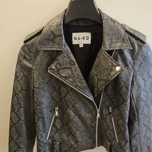 Grå ormmönstrad skinnjacka från NA-KD - Säljer en grå skinnjacka från NA-KD med ormmönster och silvriga dragkedjor. Jackan har klassisk krage, axelklaffar och två fickor med dragkedja. Perfekt för dig som vill sticka ut med en edgy stil.