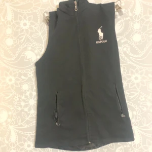 Svart väst från US Polo - Svart väst med hög krage och dragkedja framtill från US Polo. Västen har två fickor med dragkedja och en vit logotyp på bröstet. Perfekt för lager-på-lager och enkel att matcha med olika outfits.