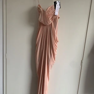 Champagnefärgad långklänning - Säljer en elegant champagnefärgad långklänning med vacker drapering och omlott-detalj upptill. Klänningen har ett snyggt fall som ger en lyxig känsla. Perfekt för speciella tillfällen! Prislappen är på!!