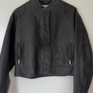 Grå skinnjacka från & Other Stories - Säljer en grå skinnjacka från & Other Stories med croppad passform. Jackan har dragkedja framtill och vid fickorna samt rund krage och markerade sömmar på ärmarna. Storlek M