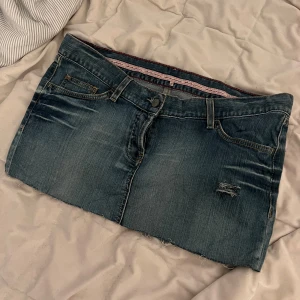 Jeanskjol med slitna detaljer - Säljer en klassisk blå jeanskjol med slitna detaljer och rå kant nertill. Kjolen har fem fickor, bälteshällor och en liten reva framtill för en cool look. Perfekt för dig som gillar en avslappnad och trendig stil.