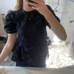 Svart blus med puffärmar - Säljer en elegant svart blus med korta puffärmar och ett diskret blommönster. Perfekt för en stilren look. Blusen har en rund hals och en lätt glansig finish som ger en lyxig känsla.