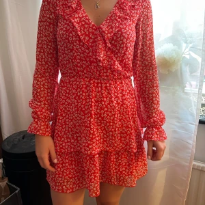 Röd blommig volangklänning - Säljer en röd kortklänning med vita småblommor och volangdetaljer. Klänningen har långärmade, lätt transparenta ärmar och v-ringning. Perfekt för vår och sommar, superfin till sneakers eller sandaler.