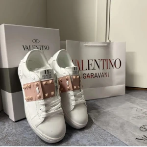Valentino Garavani Open Rosa  - Säljer ett par vita Valentino Garavani Open sneakers med ljusrosa band och guldiga nitar över vristen. Skorna har rund tå, platt sula och är tillverkade i skinn. Ikonisk design med exklusiv känsla och detaljerade nitar på sidorna. Använd fåtal gånger, inget slitage, syns bara av användning på undersidan. Boxen och påsen följer med.