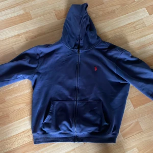 Marinblå hoodie från Polo Ralph Lauren - Säljer en marinblå hoodie från Polo Ralph Lauren med dragkedja och huva. Tröjan har två fickor framtill och en röd broderad logga på bröstet. Perfekt för en avslappnad stil och enkel att matcha med det mesta. Pris kan diskuteras