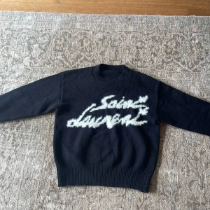 Svart stickad tröja från Saint Laurent - Säljer en svart stickad tröja från Saint Laurent med vit, fluffig logotyp framtill. Tröjan har rund halsringning och långa ärmar. Perfekt för dig som vill ha en statement piece i garderoben.