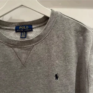 Grå sweatshirt från Polo Ralph Lauren - Säljer en klassisk grå sweatshirt från Ralph Lauren med deras ikoniska logga broderad på bröstet. Tröjan har en rund halsringning och långa ärmar, perfekt för en avslappnad stil. Passar storlek S/M. Nypris ligg på ungefär 1100kr. Inga defekter, pris ej hugget i sten.