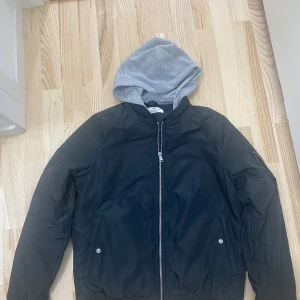 Svart bomberjacka med grå luva från H&M - Säljer en svart bomberjacka från H&M med grå huva. Jackan har dragkedja framtill, två fickor med knappar och en klassisk bombermodell. Perfekt för dig som vill ha en stilren och bekväm jacka med sportig känsla.