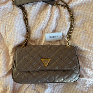 axelväska från Guess - Säljer en quiltad axelväska från Guess med guldfärgad kedjerem och klassisk trekantslogga framtill. Väskan har elegant rutmönster och passar perfekt för att lyfta din outfit. Inköpt för 1549kr, aldrig använd 😊beige/grå/lila färg. 950kr vid snabbt köp❤️