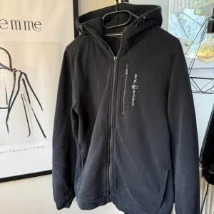 Svart Hoodie  från Sail Racing - Svart hoodiejacka från Sail Racing med saknad dragkedja framtill och färgen på bröstfickans dragkedja har tappats lite, är i storlek 170 med nån enstaka fläck vid bröstet